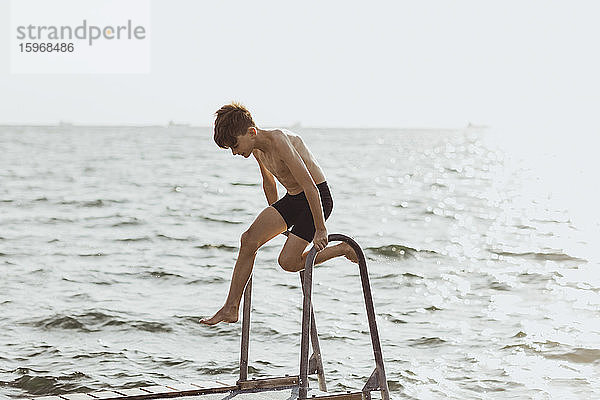 Verspielter Junge ohne Hemd  der an einem sonnigen Tag auf einer Leiter gegen das Meer balanciert