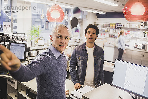 Männlicher Besitzer zeigt auf einen Kunden im Hintergrund im Elektronikladen
