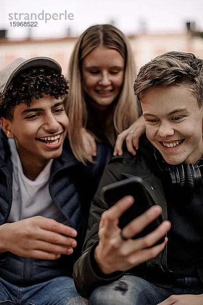 Glückliche Teenager nutzen soziale Medien auf dem Mobiltelefon  während sie in der Stadt sitzen