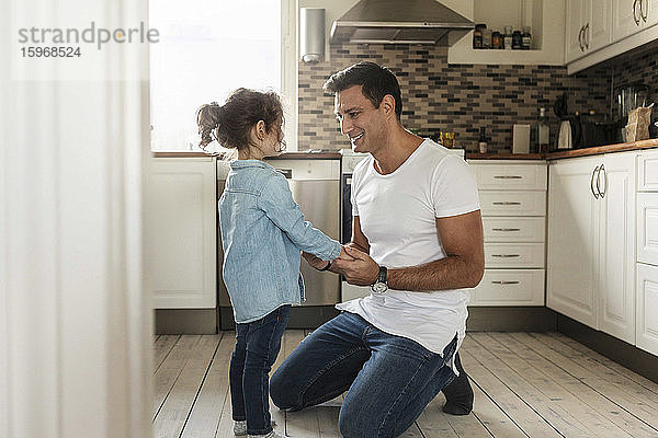 Lächelnder Vater hält mit Tochter Händchen  während er auf dem Küchenboden kniet