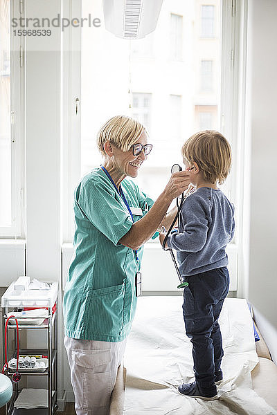 Lächelnde Ärztin hält Stethoskop an das Ohr des Jungen  während sie im Krankenhaus steht