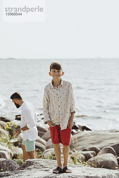 Porträt eines lächelnden Jungen auf einem Felsen gegen das Meer  während der Vater im Hintergrund steht