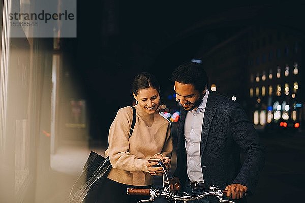 Lächelnde Frau zeigt einem männlichen Freund mit Fahrrad ein Smartphone  während sie nachts in der Stadt steht