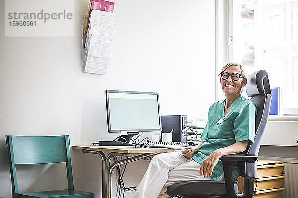 Porträt einer lächelnden  reifen Ärztin  die auf einem Stuhl am Klinikpult sitzt
