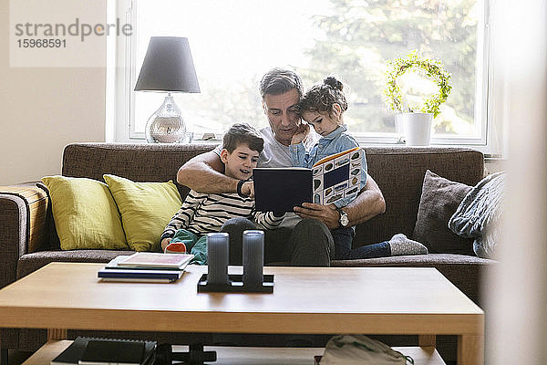 Vater liest Sohn und Tochter ein Buch vor  während sie zu Hause auf dem Sofa sitzen