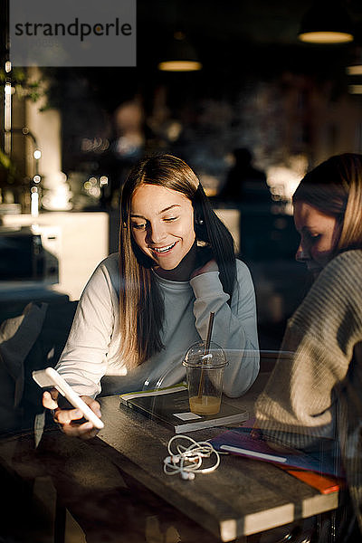 Lächelndes Teenager-Mädchen zeigt Freundin Smartphone  während sie im Café sitzt