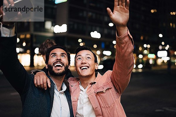 Glücklicher Mann und männlicher Freund schauen auf  während sie nachts in der Stadt gestikulieren