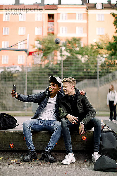 Teenager  der sich mit einem männlichen Freund per Mobiltelefon im Park sitzend selbstständig macht