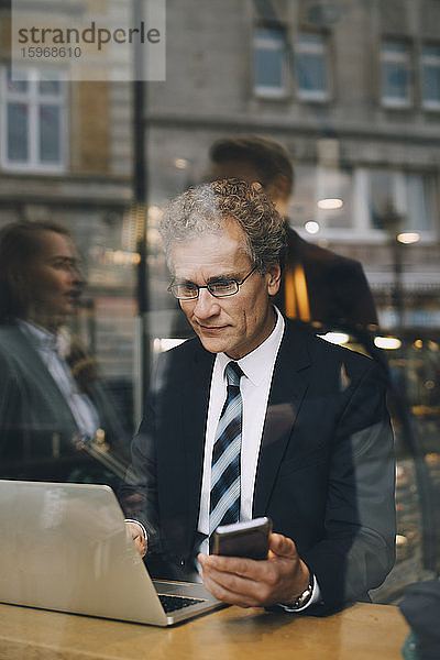 Lächelnder Geschäftsmann mit Smartphone mit Laptop im Café durch Glasfenster gesehen