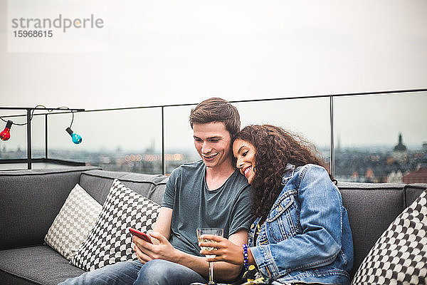 Lächelnder Mann zeigt Smartphone der Partnerin mit Getränk auf der Terrasse