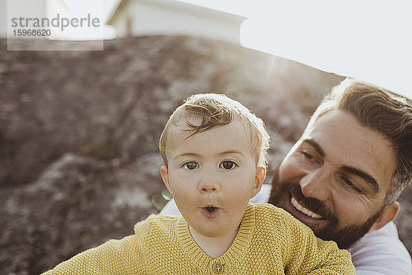 Porträt eines Kleinkindes mit schauendem und lächelndem Vater  der auf einer Felsformation sitzt