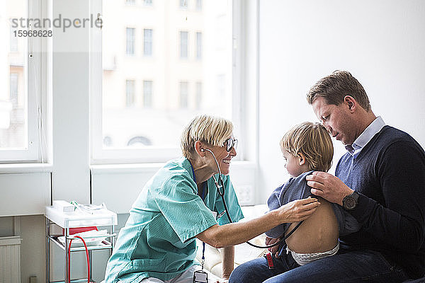 Lächelnde Ärztin untersucht den Rücken des Jungen mit dem Stethoskop  während sie im Krankenhaus beim Vater sitzt