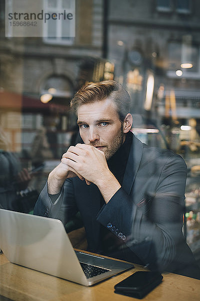 Porträt eines Geschäftsmannes mit Laptop im Café sitzend durch Glasfenster gesehen