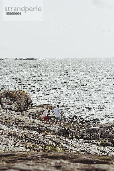 Rückansicht eines Vaters mit Sohn  der auf felsigem Land gegen den Himmel in Richtung Meer läuft