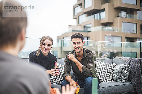 Lächelnder Mann und lächelnde Frau schauen die Freundin an  während sie während der Party auf der Gebäudeterrasse auf dem Sofa sitzen