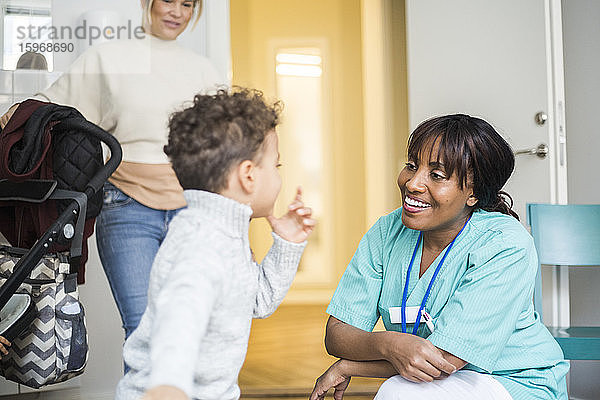 Junge spricht mit lächelnder Krankenschwester  während die Mutter in der medizinischen Klinik im Hintergrund steht