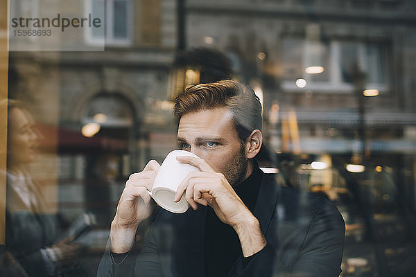 Geschäftsmann trinkt Kaffee  während er im Café durch ein Glasfenster wegschaut