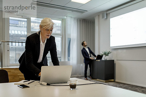 Geschäftsfrau benutzt Laptop  während ein Kollege während einer globalen Konferenzsitzung im Büro an einem Flachbildschirm steht