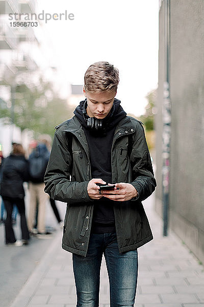 Jugendlicher Junge benutzt Mobiltelefon beim Gehen auf einem Fußweg in der Stadt