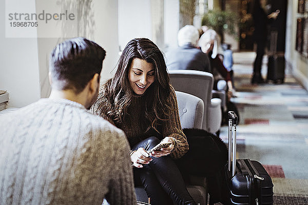 Lächelnde Frau benutzt Smartphone  während sie mit ihrem männlichen Partner auf einem Stuhl im Hotel sitzt