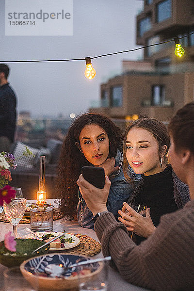 Frau zeigt Freundinnen während eines geselligen Beisammenseins auf der Terrasse des Gebäudes ihr Smartphone