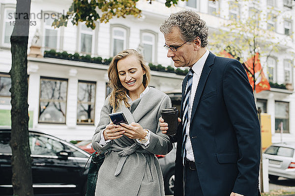 Lächelnde Geschäftsfrau zeigt einem männlichen Kollegen in der Stadt ein Smartphone