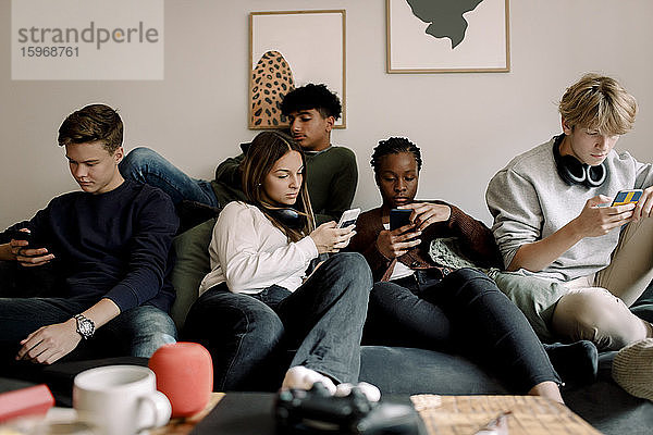 Multi-ethnische Freunde nutzen soziale Medien auf Mobiltelefonen  während sie zu Hause auf dem Sofa sitzen