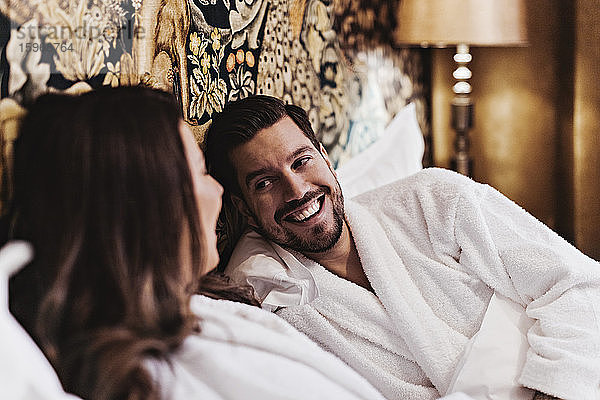 Glücklicher mittelgroßer Mann unterhält sich mit seiner Freundin  während er sich auf dem Bett im Hotelzimmer entspannt