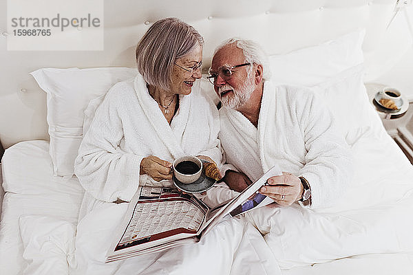Ältere Frau sieht Mann mit Zeitung in der Hand an  während sie im Hotel Kaffee auf dem Bett trinkt
