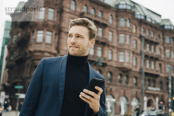 Selbstbewusster Geschäftsmann mit Smartphone schaut weg  während er sich gegen Gebäude in der Stadt stellt