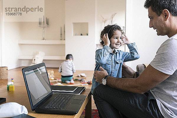 Lächelnde Tochter mit Kopfhörern steht neben dem Vater  während der Bruder zu Hause im Hintergrund spielt