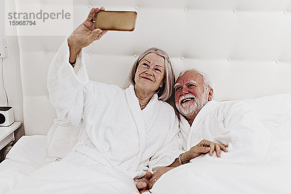 Lächelnde ältere Frau  die mit dem Mann im Hotelzimmer auf dem Bett liegend Selbstgespräche per Handy führt