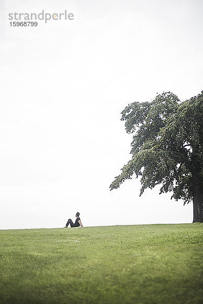 Mitteldistanzansicht eines jungen Mannes  der auf einem Grasfeld gegen den Himmel sitzt