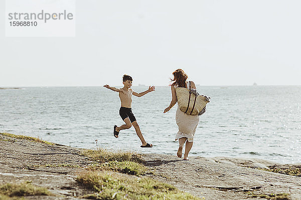 Rückansicht einer Frau mit Tasche  während ihr Sohn bei Sonnenschein gegen das Meer springt
