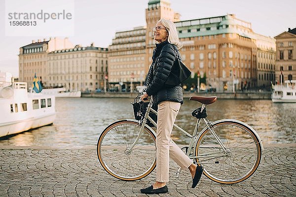 Seitenansicht einer lächelnden älteren Frau mit Fahrrad  die beim Überqueren eines Flusses in der Stadt nach oben schaut