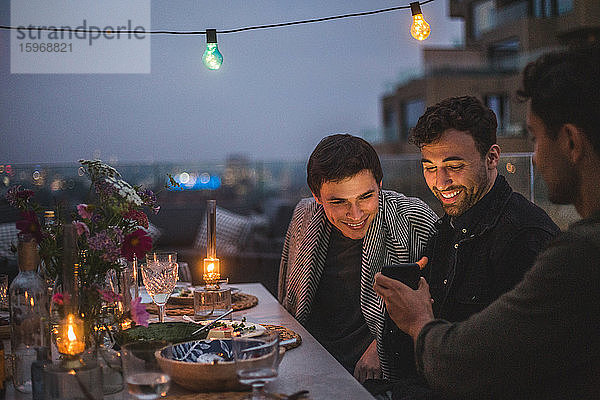 Mann zeigt lächelnden männlichen Freunden ein Smartphone  während er während des geselligen Beisammenseins auf der Terrasse sitzt