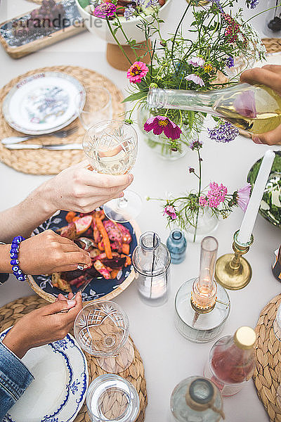 Schrägaufnahme einer geschorenen Hand  die Wein ausschenkt  während eine Frau während eines geselligen Beisammenseins auf einer Terrasse Essen serviert