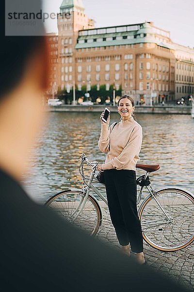 Lächelnde Frau mit Fahrrad schaut Freundin an  während sie am Fluss in der Stadt steht