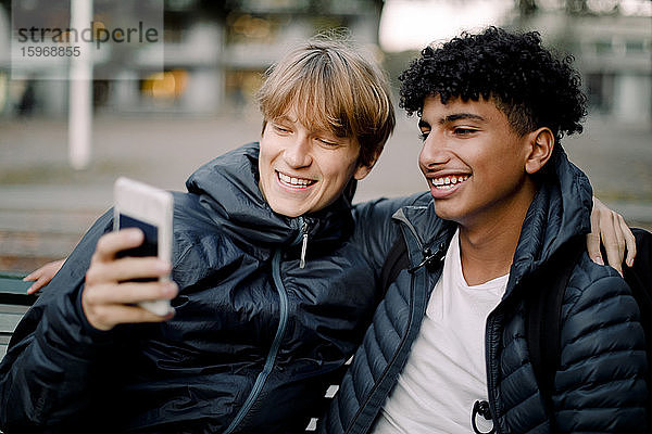 Lächelnder Teenager  der sich über ein Smartphone selbstständig macht  während er mit einem Freund in der Stadt sitzt