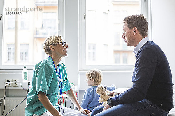 Seitenansicht einer reifen Ärztin  die mit einem Mann spricht  während ihr Sohn im Hintergrund in einer medizinischen Klinik steht