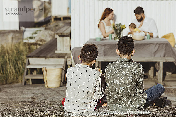 Rückansicht von männlichen Geschwistern  die über einer Decke sitzen  während die Familie mit einem kleinen Mädchen im Hintergrund