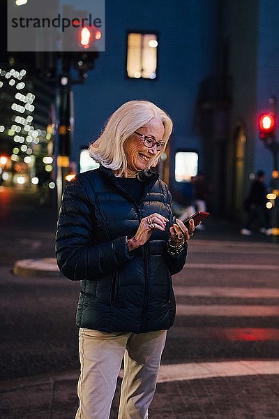 Lächelnde Frau schreibt eine SMS über ein Smartphone  während sie nachts in einer beleuchteten Stadt steht