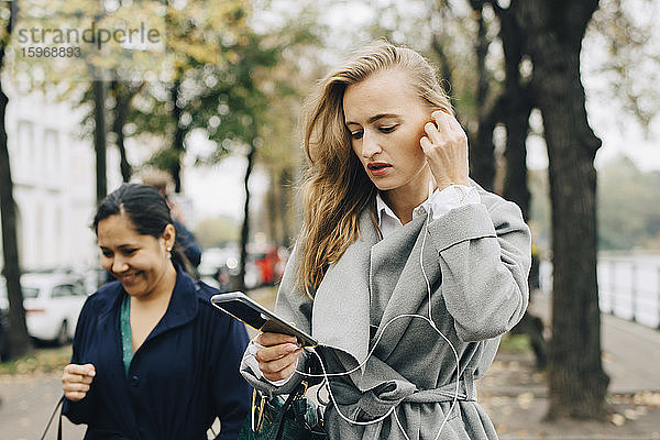 Geschäftsfrau trägt In-Ohr-Kopfhörer beim Blick auf Smartphone in der Stadt