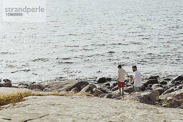 Rückansicht eines Vaters  der mit seinem Sohn Händchen hält  während er am Meer auf Felsen steht