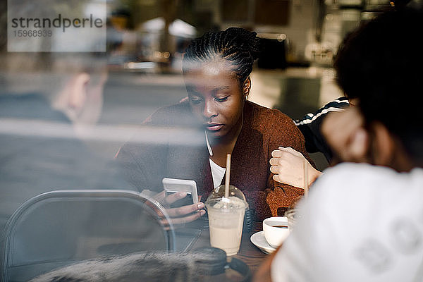 Teenager-Mädchen benutzt Mobiltelefon  während sie mit Freunden im Café sitzt  durch Glasfenster gesehen