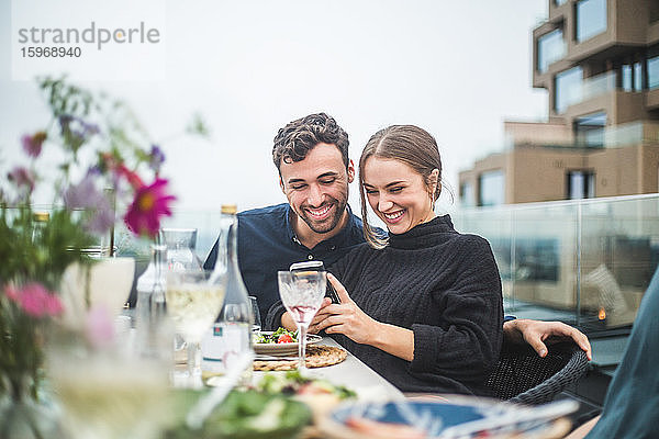 Glückliche männliche und weibliche Freunde verwenden ein Telefonfoto während einer Dachfeier