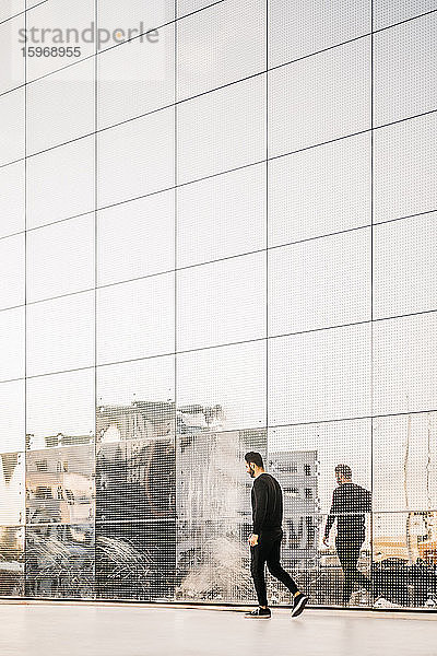 Ganzflächige Seitenansicht eines jungen Mannes  der durch Reflexion an einem Glasgebäude geht