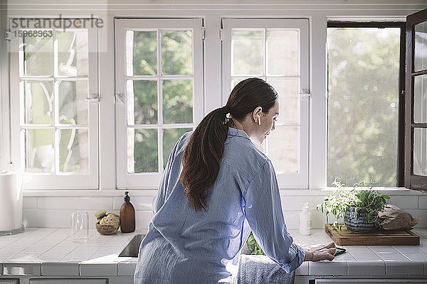 Rückansicht einer Geschäftsfrau  die ein Smartphone auf der Küchentheke im Heimbüro benutzt