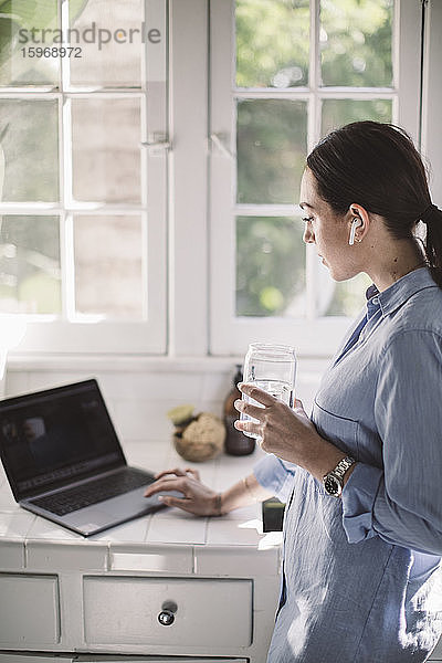 Seitenansicht eines weiblichen Mitarbeiters  der seinen Laptop am Tresen benutzt  während er im Heimbüro Wasser trinkt