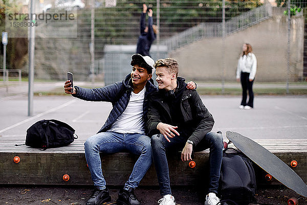 Teenager-Jungen in voller Länge  die mit dem Handy Selfie machen  während sie im Park sitzen